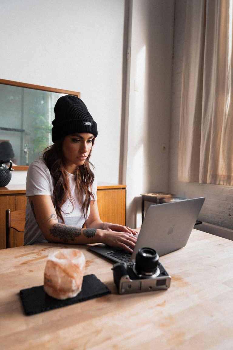 Female photographer looks at laptop while typing looking concentrated, camera lying on table