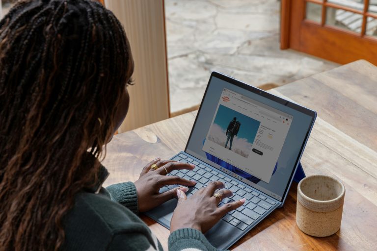 Woman with braided hair sits at a table with coffee mug while typing on a laptop showing a web search