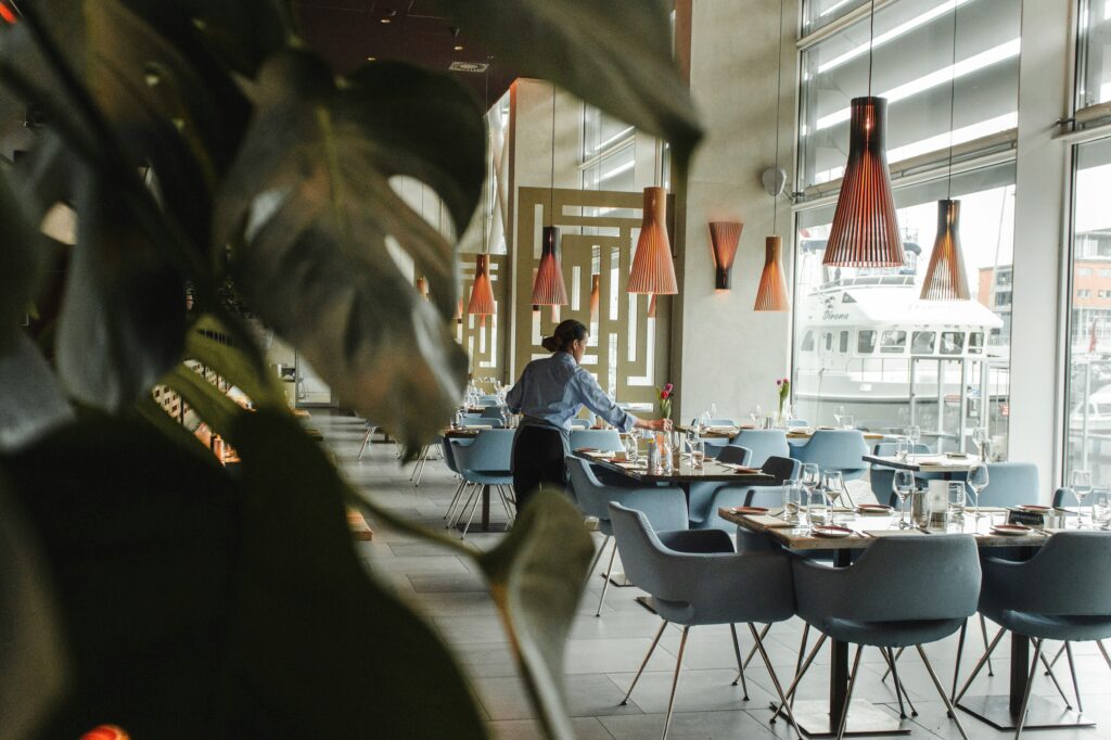 Server setting tables in a restaurant with windows overlooking a harbor.