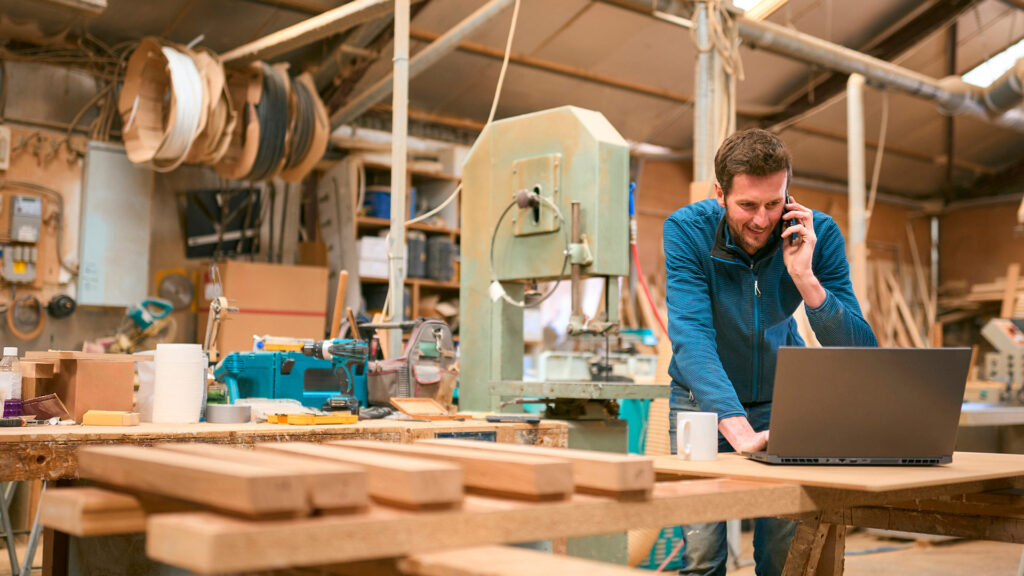 Carpenter in workshop working on laptop and cell phone.