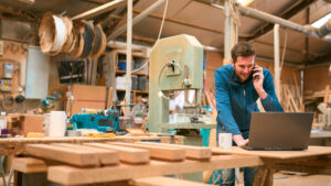 Carpenter in workshop working on laptop and cell phone.