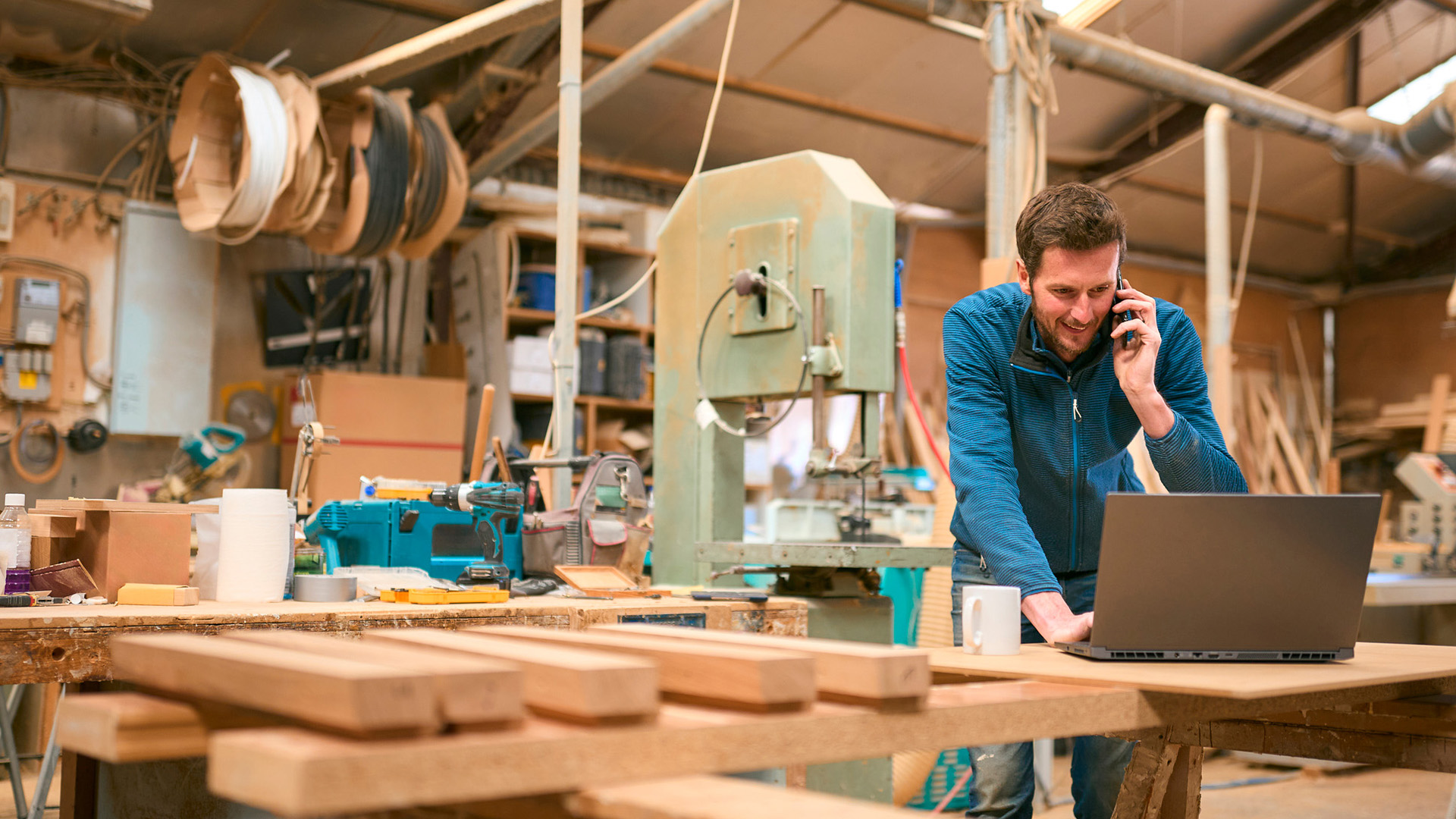 Carpenter in workshop working on laptop and cell phone.