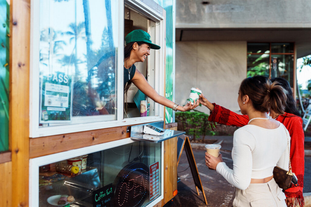 A smiling barista leaning out a coffee stand window and handing coffee to customers.