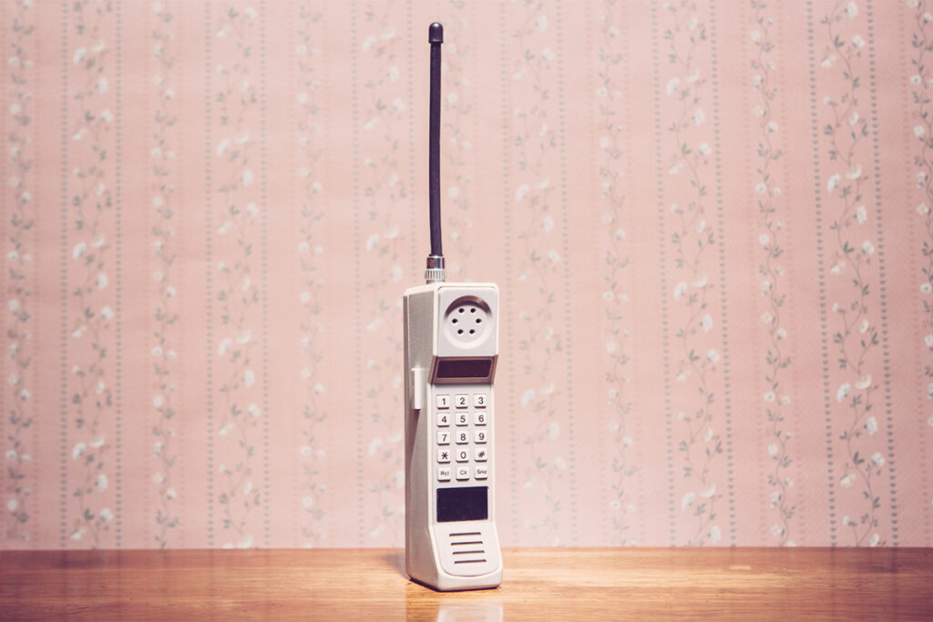 1980s or 1990s brick cell phone on a wood table with pink, flowered wallpaper behind it.
