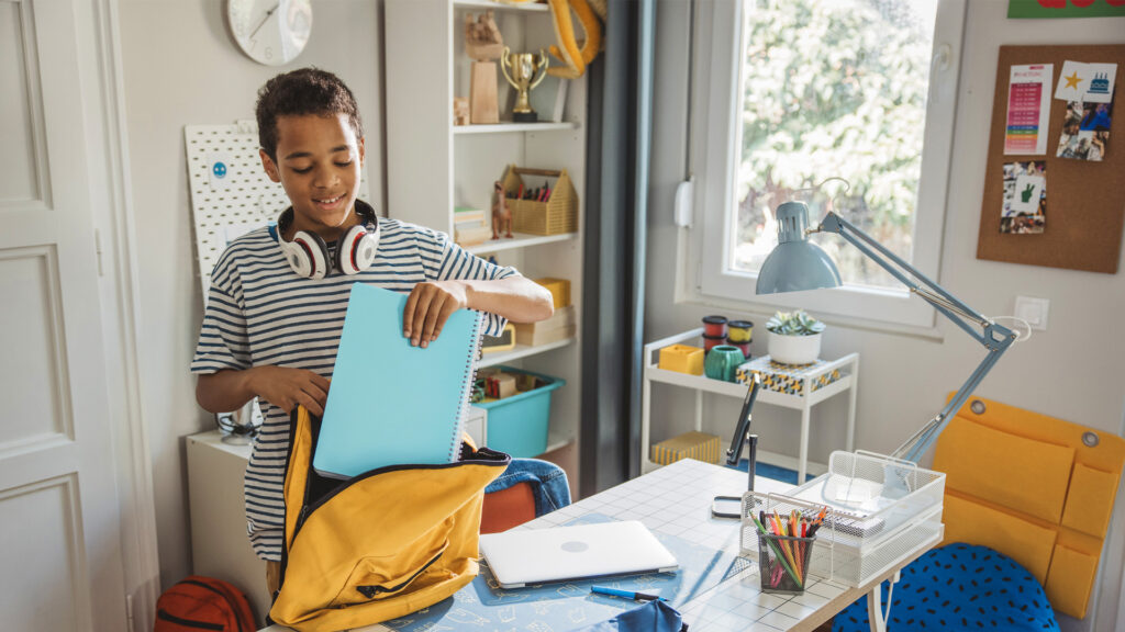 Teenage boy studding at home. He is packing backpack for school.