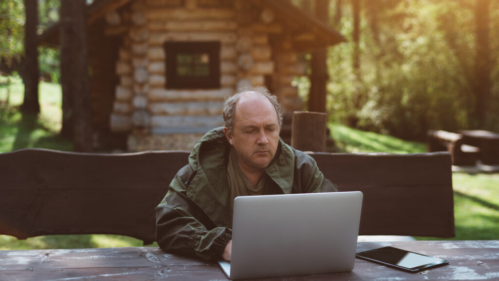 Man using a laptop in front of a cabin in the woods.
