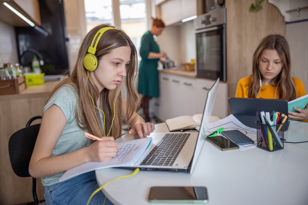 Teenage girl sitting at the table at home with her sister, attending online classes on portable information devices, mother in the kitchen in blurred background