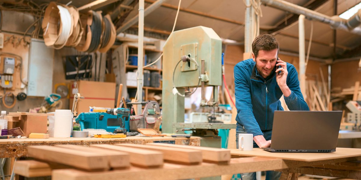 Carpenter in workshop working on laptop and cell phone.