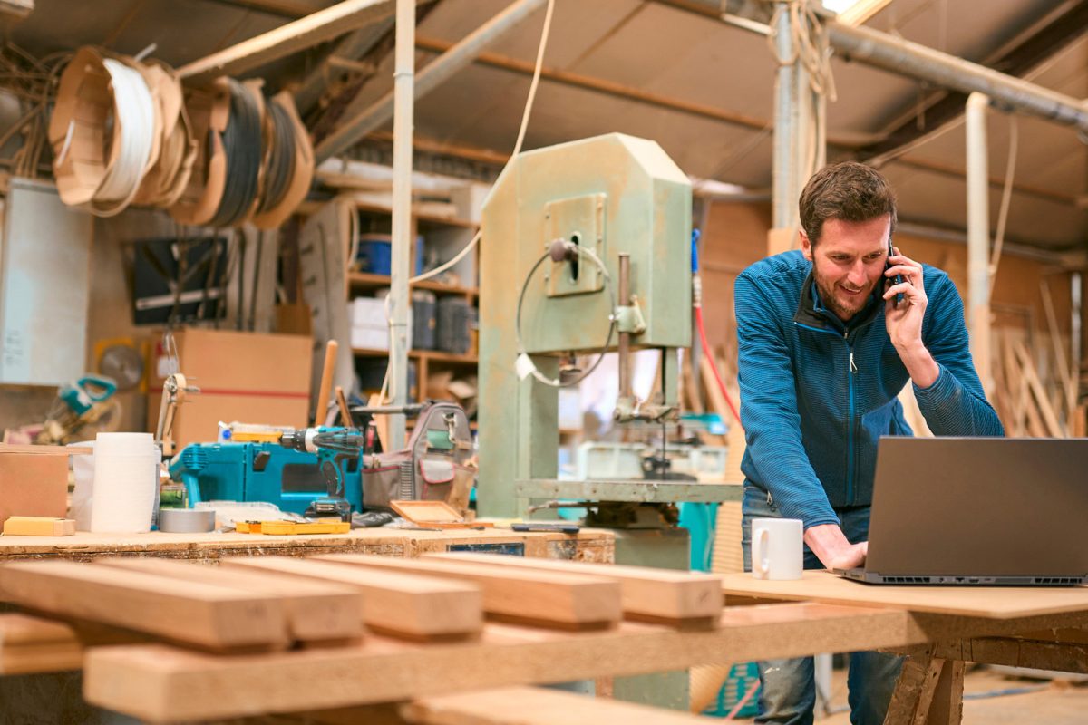 Carpenter in workshop working on laptop and cell phone.