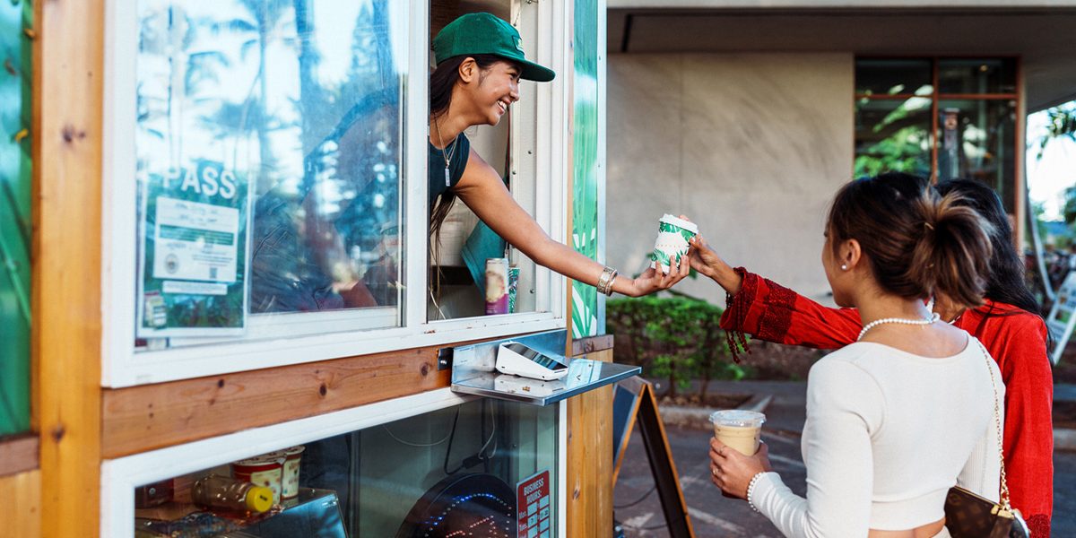 A smiling barista leaning out a coffee stand window and handing coffee to customers.