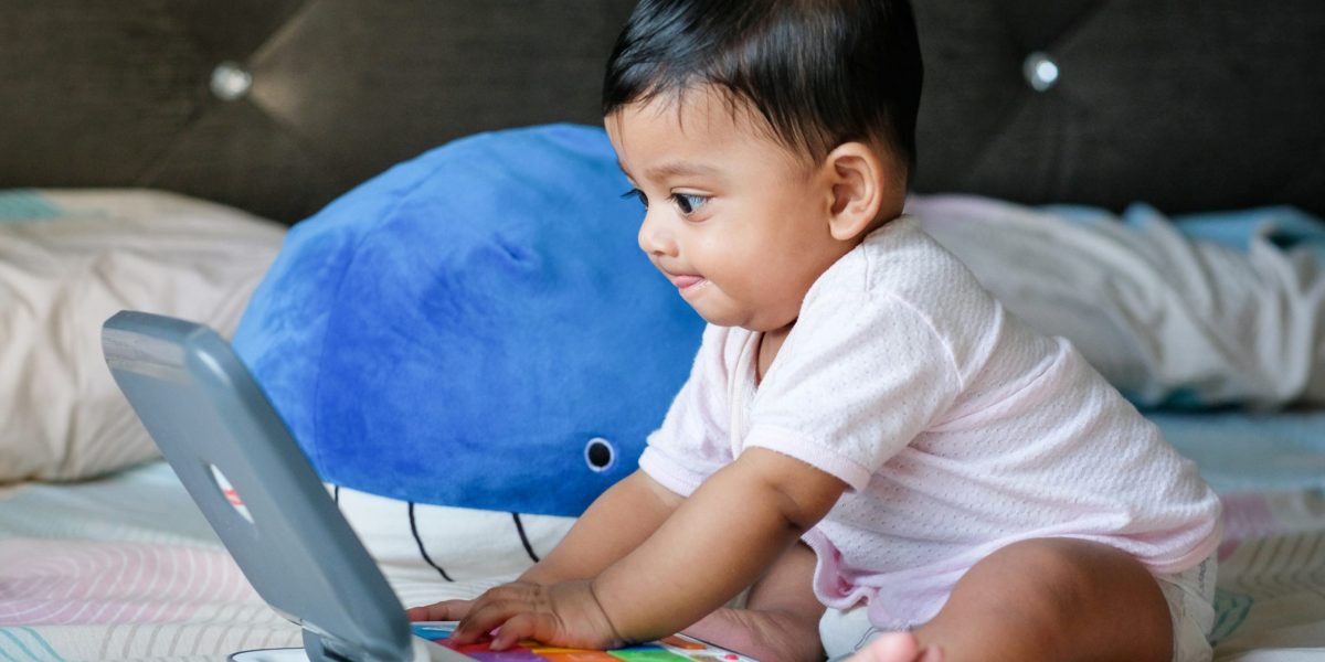 Baby with dark black hair looks focused on toy computer screen while pressing buttons, sitting next to a whale plushie